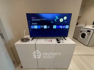 Modern living room with sleek TV setup and compact kitchen area featuring washing machine.