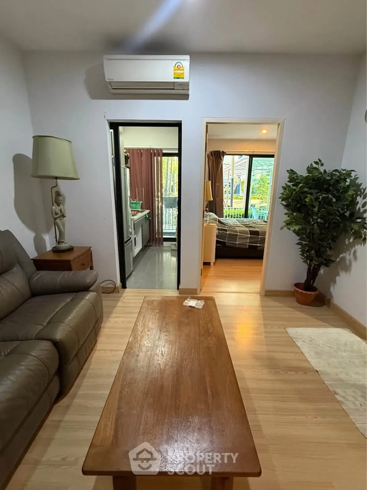 Cozy living room with wooden flooring, leather sofa, and air conditioning, leading to a modern kitchen and bedroom.