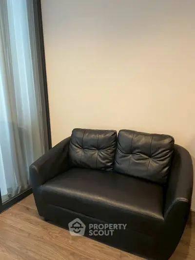 Cozy black leather loveseat in modern living room with wooden flooring and large window.