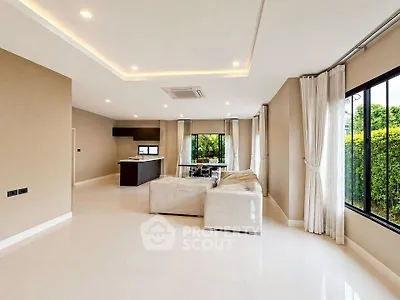 Spacious modern living room with open kitchen and large windows overlooking lush greenery.