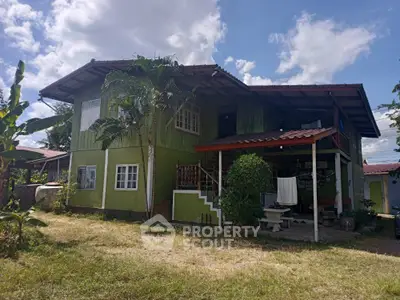 Charming two-story green home with spacious porch and lush surroundings, perfect for serene living and outdoor relaxation.