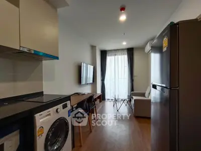 Modern living room with kitchen featuring washing machine and fridge, stylish furniture, and natural light.