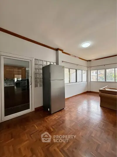Spacious living room with wooden flooring and large windows, featuring a modern refrigerator and cozy seating area.