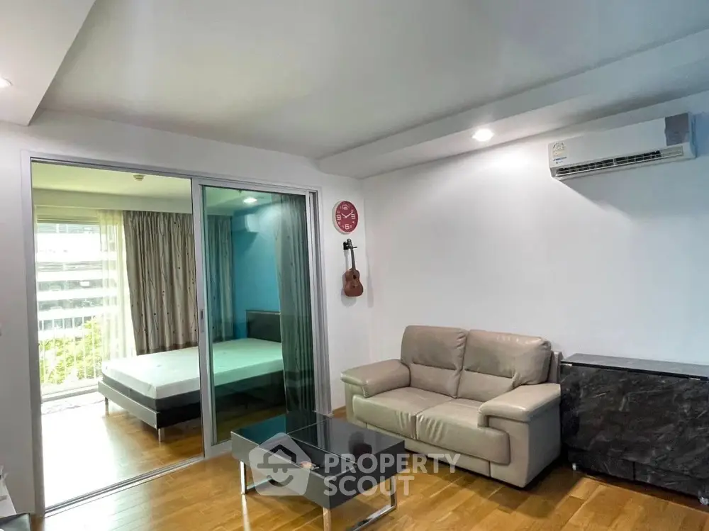 Modern living room with leather sofa and glass coffee table, adjacent to a cozy bedroom with sliding glass doors.