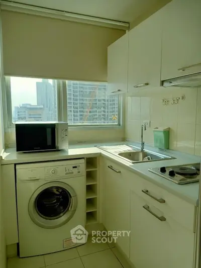 Modern kitchen with washing machine and microwave, featuring sleek white cabinetry and city view.