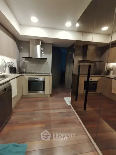 Modern kitchen with sleek cabinetry and built-in oven, featuring reflective surfaces and wooden flooring.