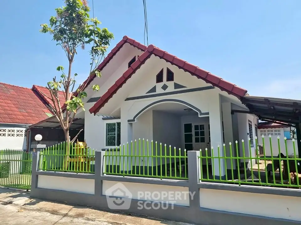 Charming single-story house with red roof and green fence in sunny neighborhood.