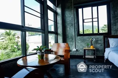 Industrial-chic bedroom with large windows offering a lush view, polished concrete walls, and stylish wooden furniture creating a modern yet cozy atmosphere.