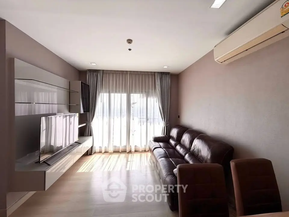 Modern living room with leather sofa, wall-mounted TV, and large windows with sheer curtains.