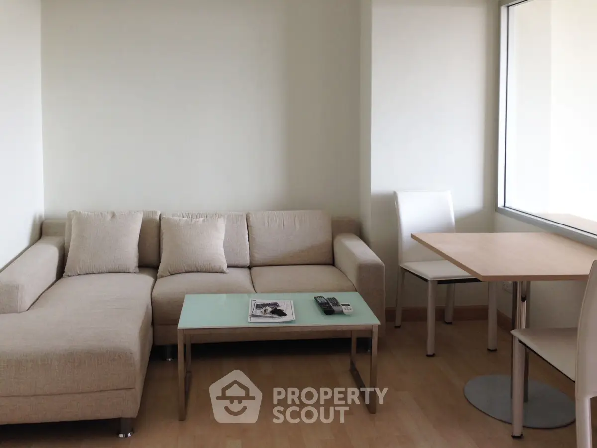 Modern living room with beige sectional sofa and glass coffee table, adjacent to a dining area with natural light.