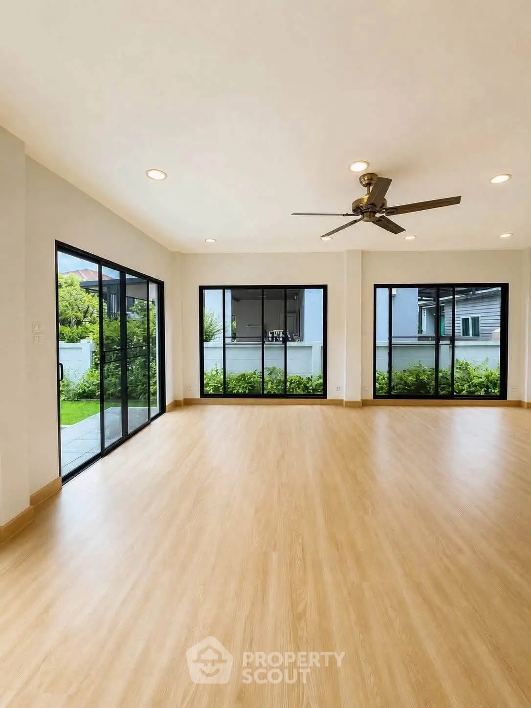 Spacious living room with large windows and garden view, featuring modern ceiling fan and wooden flooring.