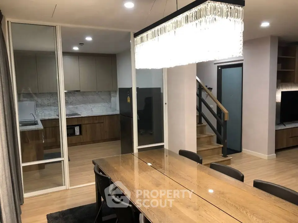 Modern dining area with elegant chandelier and view into sleek kitchen with wooden cabinetry.