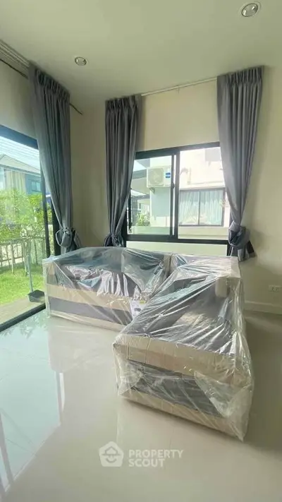 Bright living room with large windows and new furniture wrapped in plastic.