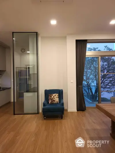 Modern living room with sleek wooden flooring, large windows offering a serene tree view, and a cozy blue armchair for relaxation.