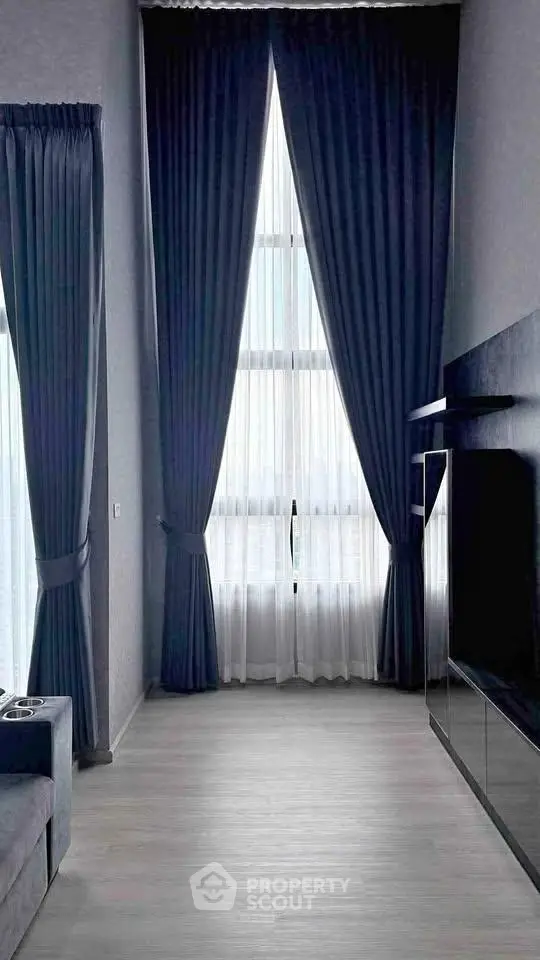 Modern living room with tall windows and elegant curtains, featuring sleek flooring and a wall-mounted TV.