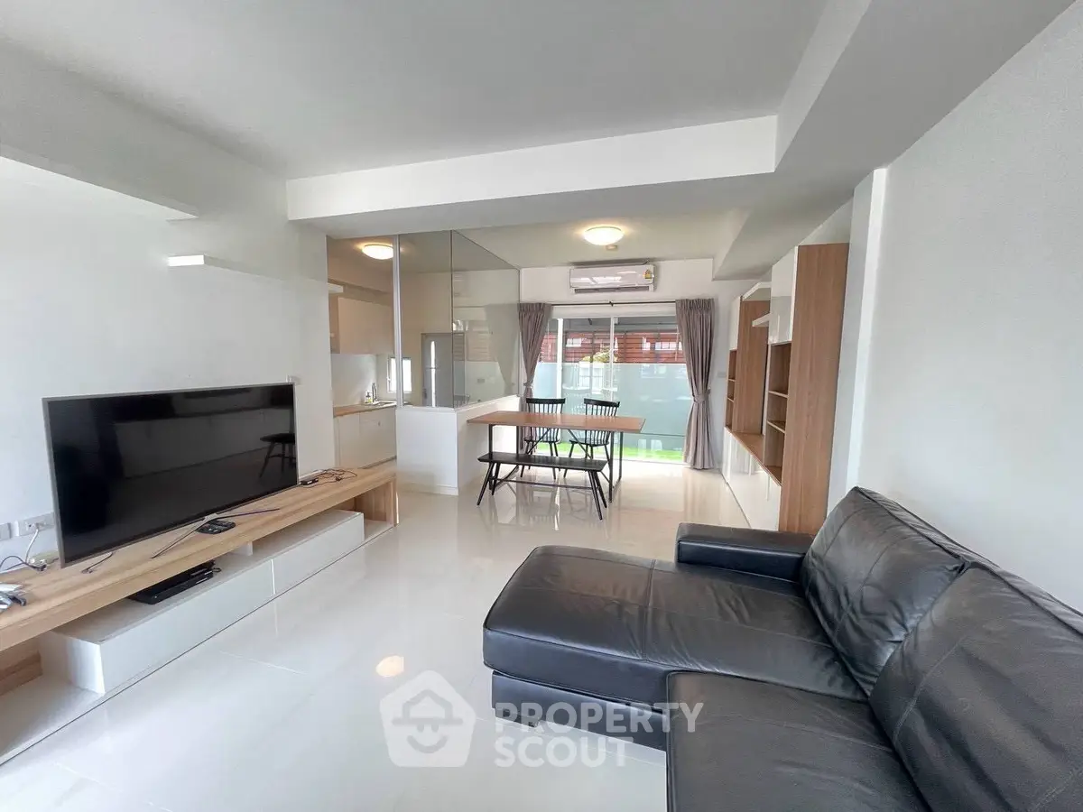 Modern living room with sleek black leather sofa and large TV, open to dining area with natural light.