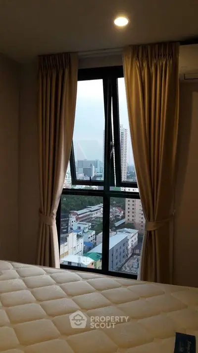 Cozy bedroom with city view through large window, featuring elegant curtains and comfortable mattress.