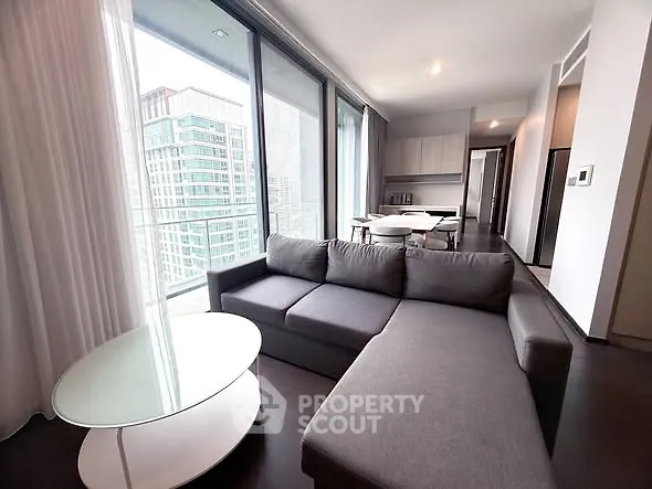 Modern living room with large windows and city view, featuring a sleek gray sofa and glass coffee table.