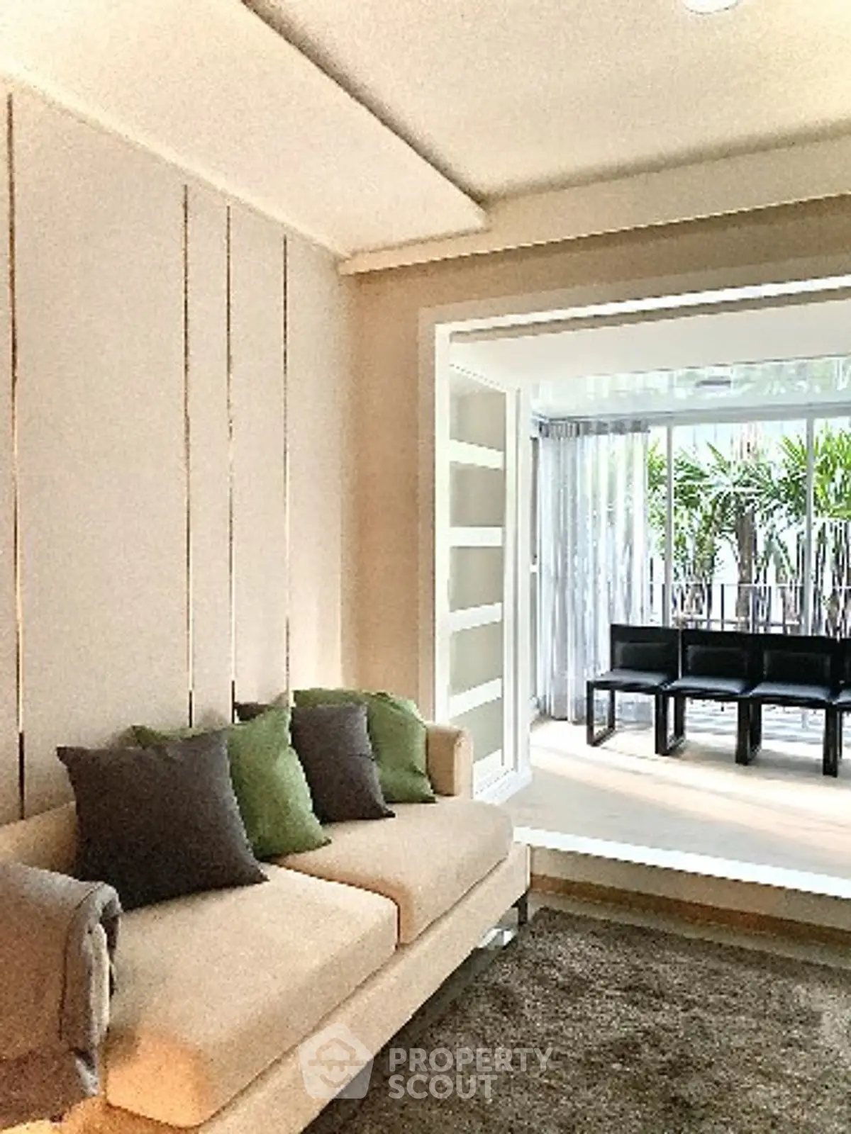 Modern living room with cozy beige sofa and green cushions, leading to a bright balcony with lush plants.