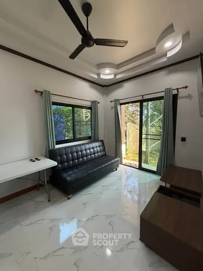 Modern living room with sleek black sofa, ceiling fan, and large windows overlooking lush greenery.
