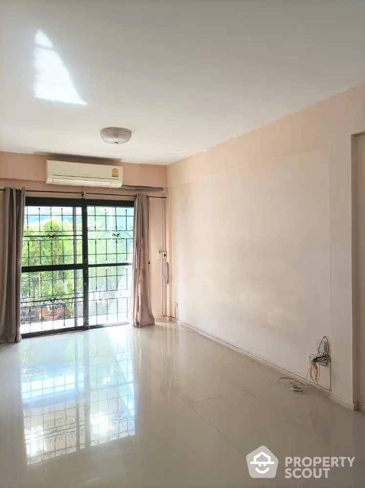 Spacious and bright living room with glossy tiled flooring, large windows leading to a balcony with a serene view, and an air conditioning unit for comfort.