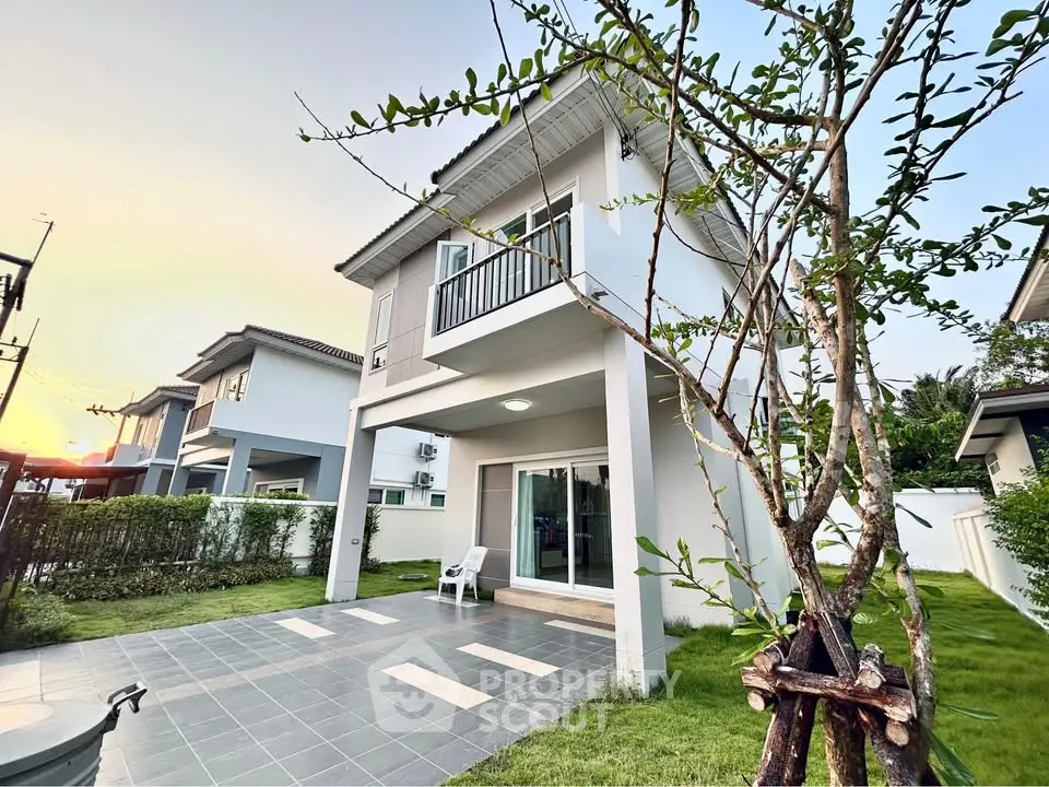 Charming two-story house with a small balcony and lush green garden at sunset.