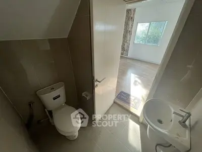Compact bathroom with toilet and sink, adjacent to a sunlit room with wooden flooring.