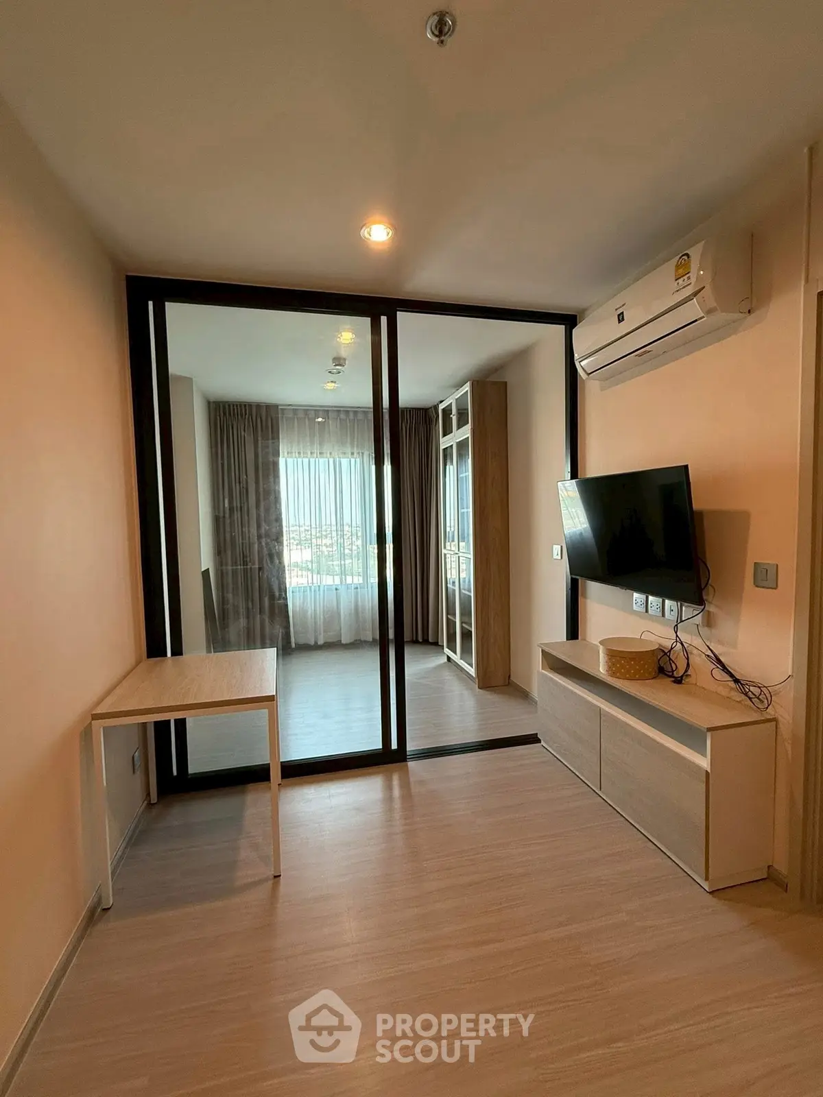 Modern living room with glass partition and wall-mounted TV, featuring sleek wooden furniture and air conditioning.