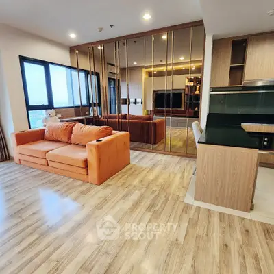 Spacious modern living room with orange sofa and mirrored wall, open to sleek kitchen area.