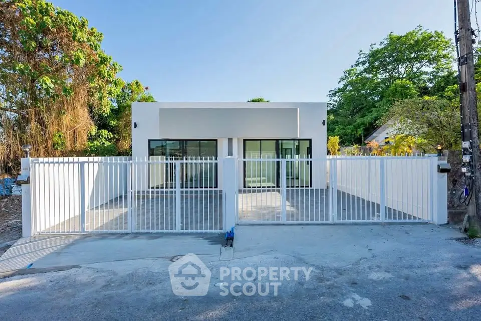 Modern minimalist house with white fence and large windows, surrounded by lush greenery.