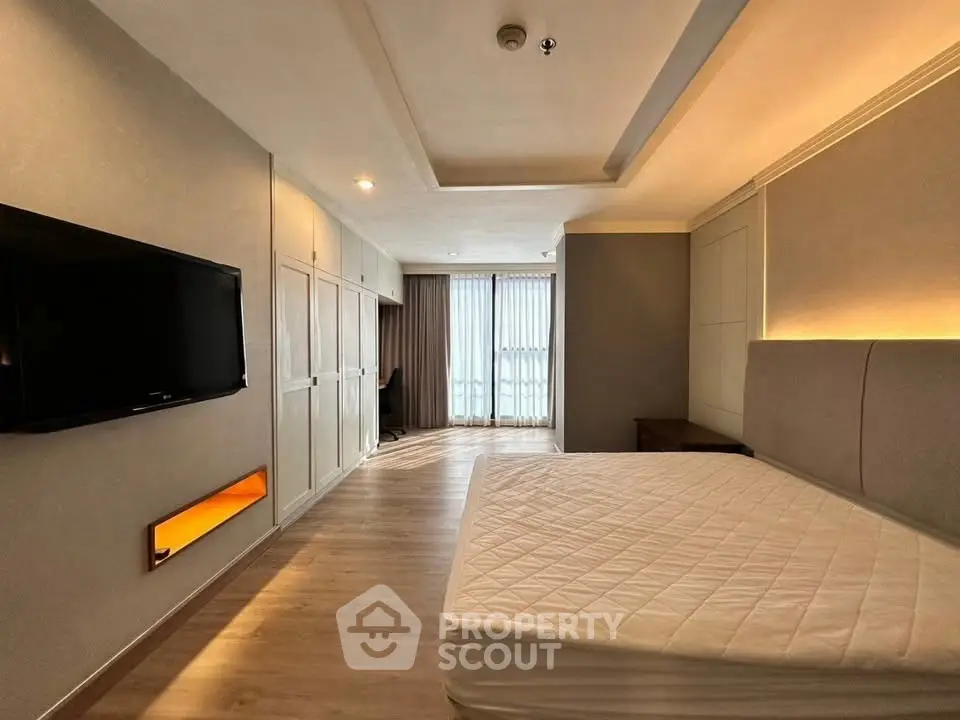 Spacious bedroom with modern design and natural light, featuring a wall-mounted TV and large windows.