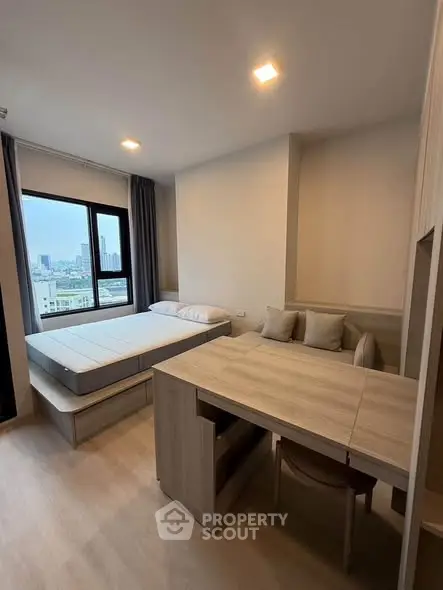 Modern studio apartment with bed and dining area, featuring large window with city view.