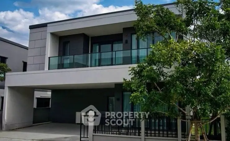 Modern two-story house with sleek design and large balcony, surrounded by lush greenery.