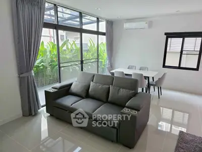Modern living room with sleek gray sofa and dining area, large windows overlooking lush greenery.