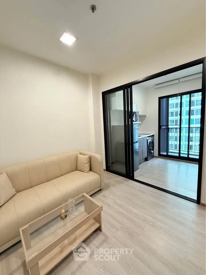 Modern living room with beige sofa and glass coffee table, adjacent to a kitchen with balcony view.