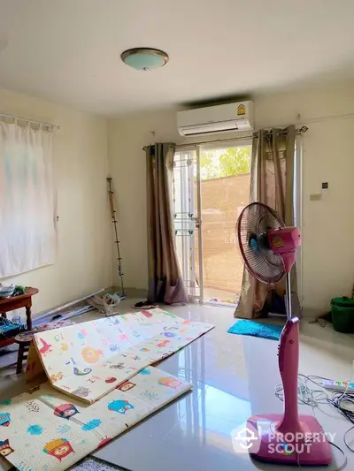 Bright and airy living space with ample natural light, featuring a large window with curtains, an air conditioning unit, and a playful children's play mat.