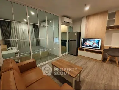 Modern living room with open kitchen and glass-walled bedroom, featuring sleek furniture and wood flooring.