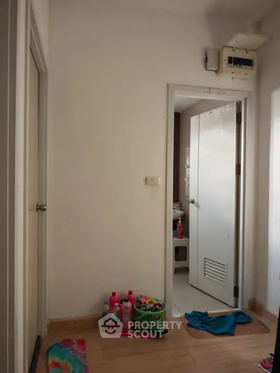 Simple hallway with open door leading to a bathroom, featuring wooden flooring and cleaning supplies.