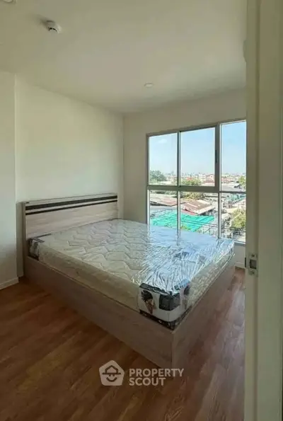 Bright bedroom with large window and city view, featuring a new mattress on a wooden floor.