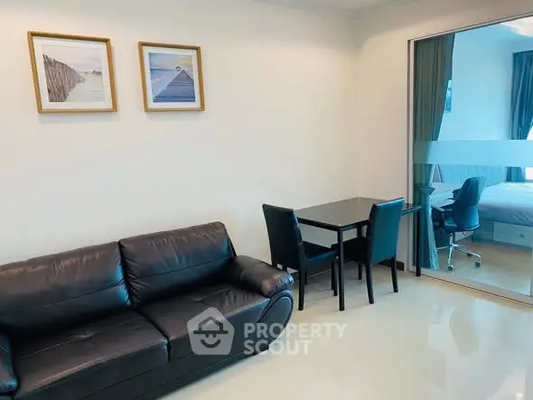 Modern living room with sleek black leather sofa and dining area, adjacent to a glass-partitioned bedroom.
