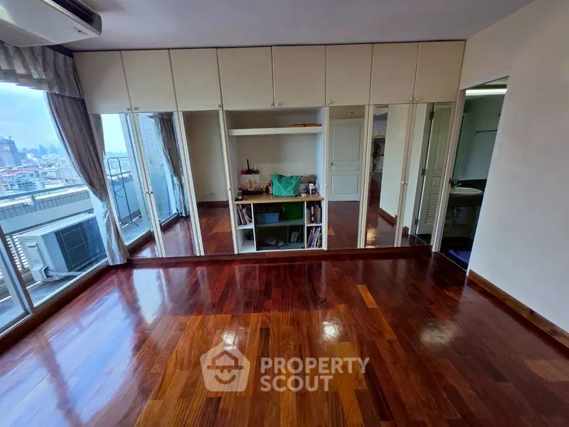 Spacious room with polished wooden floors and built-in storage, offering city views from large windows.