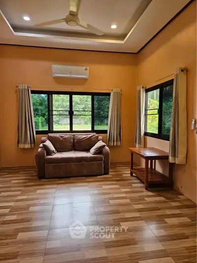 Cozy living room with large windows, air conditioning, and comfortable seating, offering a serene view of lush greenery.