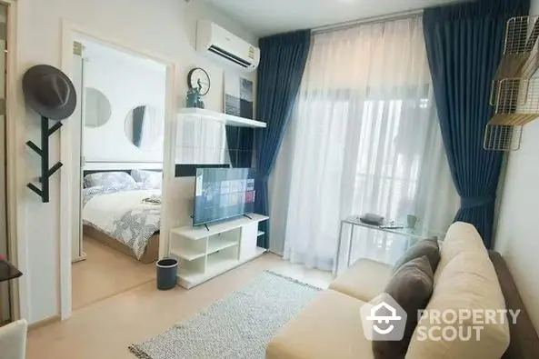 Cozy studio apartment with integrated living and sleeping area, modern furnishings, and ample natural light from large window with sheer curtains.