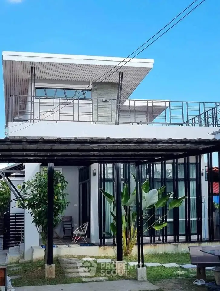 Modern two-story house with sleek design and spacious balcony, surrounded by lush greenery and clear blue sky.