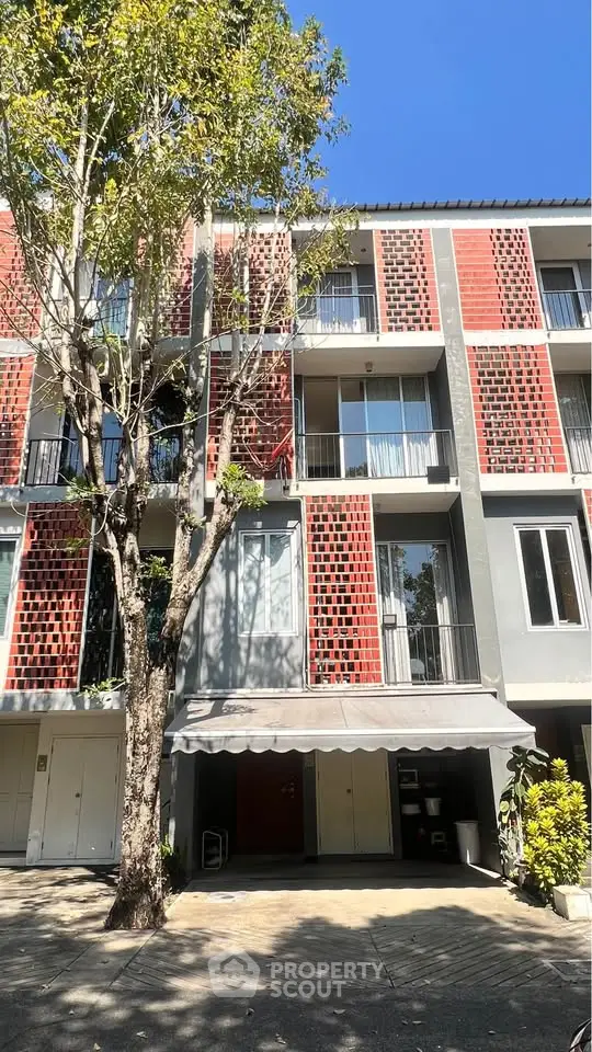 Modern multi-story building with unique brick facade and balconies, surrounded by lush greenery.