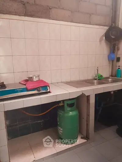 Simple kitchen with gas stove and tiled countertops, featuring a double sink and exposed brick wall.