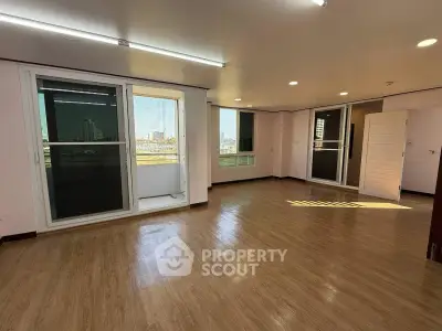 Spacious living room with large windows and balcony access, featuring sleek wooden flooring and abundant natural light.