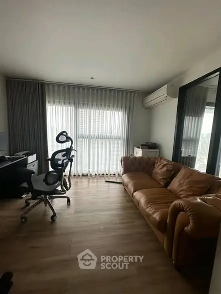 Modern living room with leather sofa and office setup, featuring large window and wooden flooring.