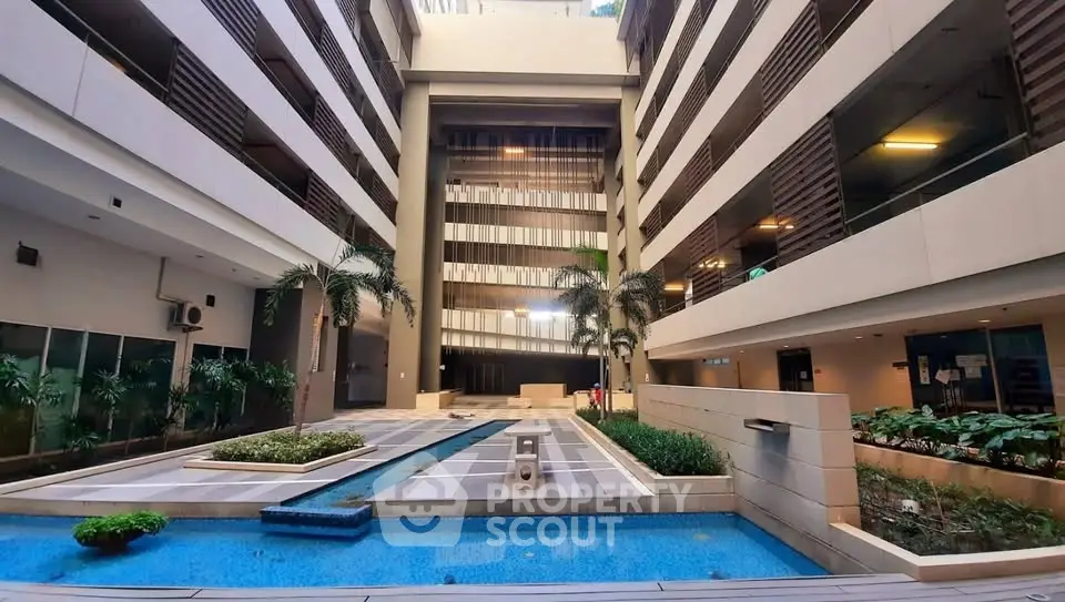 Modern apartment building with central courtyard and water feature, surrounded by lush greenery and palm trees.
