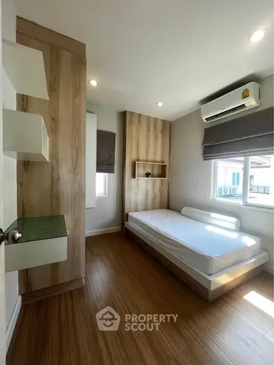 Modern bedroom with wooden flooring and natural light, featuring a minimalist design and air conditioning.
